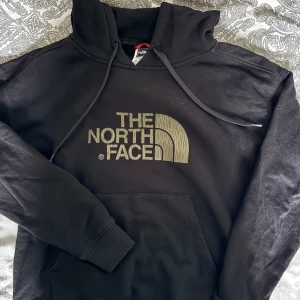 Svart hoodie från The North Face - Svart hoodie från The North Face med stor logga i beige framtill. Klassisk känguruficka och ribbade muddar. Perfekt för chill dagar eller när du vill ha en clean streetstyle-look. 