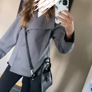 Grå, kort kappa med bred krage - Snygg grå rock med bred krage och stora knappar framtill. Jackan har en rak och något loose passform, perfekt för dig som gillar stilren och enkel design. Passar till många olika outfits och har långa ärmar.