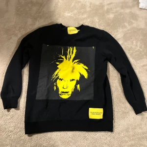 Svart Calvin Klein x Andy Warhol tröja - Svart sweatshirt från Calvin Klein Jeans Andy Warhol-kollektionen med en stor gul grafisk print av Andy Warhols ansikte på framsidan. Tröjan har rund hals, långa ärmar. Passar en 170