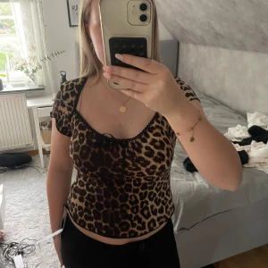 Leopardmönstrad topp med rosett - Säljer en topp med leopardmönster i brunt och beige. Toppen har korta ärmar, liten rosett framtill och svarta spetsdetaljer längs urringningen. Materialet är stretchigt och lätt, perfekt för en trendig look.