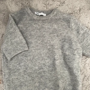 Grå stickad tröja från Zara - Mysig grå stickad tröja från Zara med rund halsringning och ribbade muddar. Tröjan har kortare ärmar och är perfekt för lager-på-lager under hösten eller vintern. Enkel och stilren design som passar till det mesta.