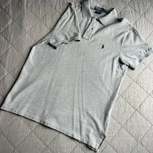 Grå pikétröja från Polo Ralph Lauren - Snygg grå pikétröja från Polo Ralph Lauren med klassisk krage och två knappar framtill. Tröjan har korta ärmar och är tillverkad i mjuk bomull. På bröstet finns den broderade Polo-loggan i vinrött.