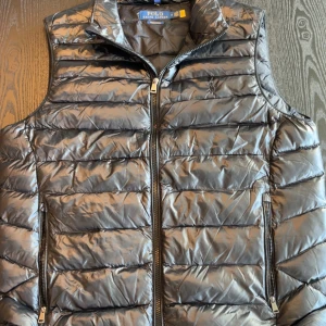 Svart dunväst från Polo Ralph Lauren - Snygg svart dunväst från Polo Ralph Lauren med quiltad design och broderad logga på bröstet. Västen har dragkedja framtill och två fickor med dragkedja. Perfekt för lager-på-lager och ger en clean look.