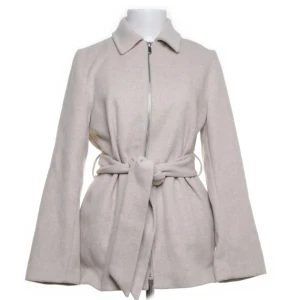 Beige jacka med bälte från Dobber XS - Stilren beige jacka från Dobber i storlek XS. Jackan har klassisk krage, dragkedja framtill och ett brett bälte i midjan för snygg passform. Långärmad och i mjukt material, perfekt för höst och vår. Enkel och clean look som passar till det mesta. 