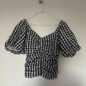 Svartvit rutig blus med puffärm - Supersöt blus med svartvitt rutigt mönster och puffiga korta ärmar. Toppen har smockad effekt och rynk framtill som ger en snygg passform. Perfekt för dig som gillar trendiga detaljer och vill sticka ut lite extra.