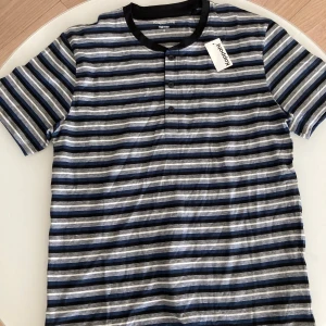 Randig t-shirt från Kappahl, blå/svart - Randig t-shirt från Kappahl med blå, svarta och vita horisontella ränder. Rund halsringning med svart kant och tre knappar framtill. Tillverkad i mjuk bomull, perfekt för en chill look.