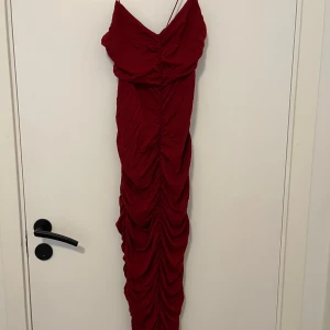 Röd långklänning från ASOS DESIGN - Supersnygg röd långklänning från ASOS DESIGN med smala axelband och draperad, rynkad design längs hela klänningen. Figurnära passform som framhäver siluetten och ger en elegant look. Perfekt för dig som vill sticka ut med en djärv färg och trendig stil.