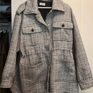 Grå jacka från Cloeys - Snygg och trendig grå overshirt från Cloeys med svartvitt rutigt mönster. Jackan har stora bröstfickor med knappar, markerad krage och tryckknappar framtill. Oversized passform och långa ärmar, perfekt för lager-på-lager-stil.