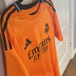Real Madrid Valverde Adidas tröja M - Säljer en orange Real Madrid fotbollströja från Adidas med Valverde och nummer 8 på ryggen. Tröjan har svarta detaljer, korta ärmar och är tillverkad i ett lätt, ventilerande funktionsmaterial. Klassisk Adidas-design med tre svarta ränder på axlarna och klubbmärke på bröstet.