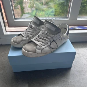 Philippe Model Junior PRSX - Snygga vita sneakers från Philippe Model Junior med grå mockadetaljer och klassisk snörning. Skorna har en rund tå, platt sula och ikonisk sköldlogga på sidan. Tillverkade i skinn och mocka för en lyxig känsla. Perfekta för dig som gillar stilrena och trendiga sneakers.