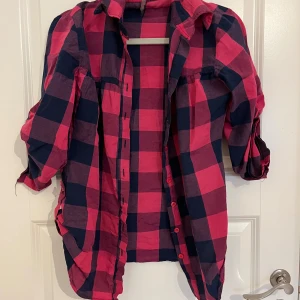 Rutig rosa och mörkblå skjorta Red Queen - Snygg rutig skjorta från Red Queen i storlek M. Passar L oxå.  Skjortan har ett mönster i rosa och mörkblått, klassisk krage och knappar framtill. Ärmarna är långa men kan rullas upp och fästas med knapp. Perfekt för dig som gillar färg och mönster.