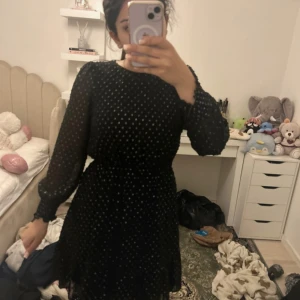 Svart kortklänning med glitterprickar - Supersöt svart kortklänning med långa ärmar och glittriga silverprickar. Klänningen har en lätt transparent känsla i ärmarna och markerad midja för en snygg siluett. Perfekt för dig som vill sticka ut lite extra! Materialet känns lätt och flowy.