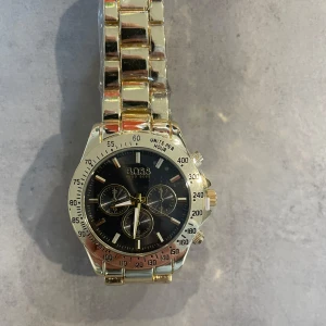 Guldfärgad klocka från BOSS - Stilren klocka från BOSS med guldfärgad länk och svart urtavla. Klockan har tre mindre urtavlor, tydliga markeringar och kronograf-funktion. Metallarmbandet har en blank finish och BOSS-logga på spännet.