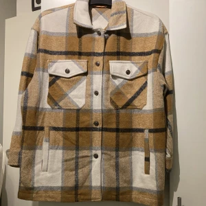 Rutig fleecejacka / overshirt för dam - Fleecejacka / overshirt för dam i rutigt mönster. Jackan har bruna, beige och vita toner med svarta och grå ränder, två bröstfickor med lock och knappar samt sköna sidofickor att stoppa händerna i. Materialet är mjukt och ulligt. Ofodrad. Passar bra för lager-på-lager-stil. 