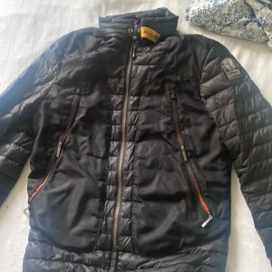 Svart quiltad pufferjacka Parajumpers - Svart pufferjacka från Parajumpers med quiltad design, hög krage med gul detalj och patch på ärmen. Jackan har två snedställda fickor med dragkedja och markerade sömmar på ryggen. Perfekt för kyliga dagar och riktigt snygg streetstil.