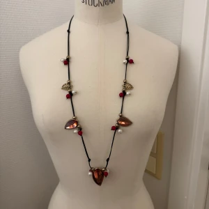 Långt halsband med röda och pärlvita detaljer - Unikt långt halsband med svarta band, stora droppformade röda stenar, pärlvita och röda pärlor samt små guldfärgade löv. Lättmetall/ plast. Krimskrams. GRATIS om du köper något annat av mig. Hämtas i Hägersten om du vill slippa frakt. Var även uppmärksam på när Plick erbjuder fraktfritt ibland på dyrare plagg. Följ mig gärna. Har mycket att lägga ut i olika storlekar! 