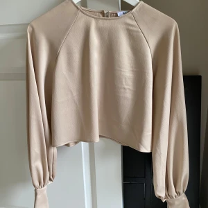 Beige croppad blus med vida ärmar - Säljer en stilren beige blus från NA-KD med croppad passform och långa, vida ärmar med breda muddar.                                               Blusen  har rund halsringning och är tillverkad i ett tungt och silkigt material som ger ett snyggt fall. 