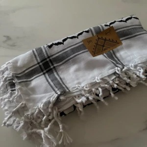 Vit och svart palestinasjal med tofsar - Säljer en klassisk palestinasjal i vitt och svart med snyggt rutigt och prickigt mönster. Sjalen har dekorativa tofsar längs kanterna och är tillverkad i mjukt bomullsmaterial. Perfekt att ha runt halsen eller som accessoar. Stor och fyrkantig form.