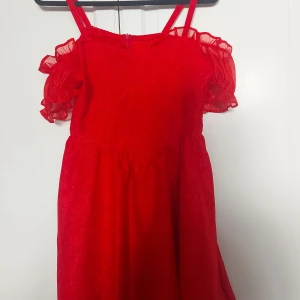 Röd axelbandsklänning med volang - ⚠️ Skriv till mig innan köp⚠️.                                        Har ett hål i tyget! Se sista bilden.                                            Superfin röd kortklänning med axelband och volangiga offshoulder-ärmar. Klänningen har blommigt mönster i tyget och är gjord i ett glansigt material som känns lyxigt. Dragkedja bak för enkel påklädning. Perfekt för dig som vill sticka ut!