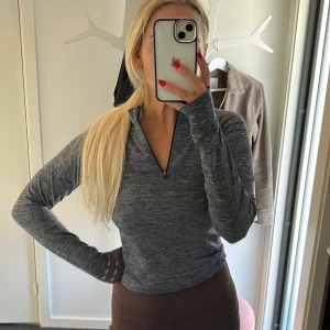 Grå långärmad träningströja half zip - Superskön grå långärmad träningströja med half zip och coola håldetaljer vid ärmsluten. Tröjan har en slim passform och är gjord i ett mjukt, stretchigt syntetmaterial som andas. Perfekt för gymmet eller löprundan!