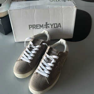 Beige sneakers från Premiata - Snygga beige sneakers från Premiata med vit hälkappa och vita skosnören. Skorna har perforerad tå, svart yttersula och diskret logga på sidan. Tillverkade i mocka och skinn för en clean och stilren look. Perfekta för dig som gillar minimalistisk stil. Kan tyvärr inte garantera äkta, köpta på Vinted för 1299