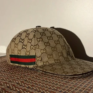Klassisk beige Gucci keps - Snygg keps från Gucci i beige med klassiskt GG-monogram över hela kepsen. Har en ikonisk röd och grön rand på sidan och justerbart spänne bak i brunt läder. Tillverkad i bomullscanvas med detaljer i läder. Perfekt för dig som vill sticka ut med en lyxig accessoar.