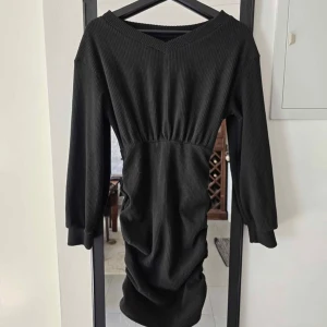 Svart ribbad långärmad klänning - Svart ribbad klänning med v-ringning och långa ärmar. Modellen är figurnära med markerad midja och har en mjuk, stretchig känsla. Perfekt för dig som gillar en enkel men stilren look.