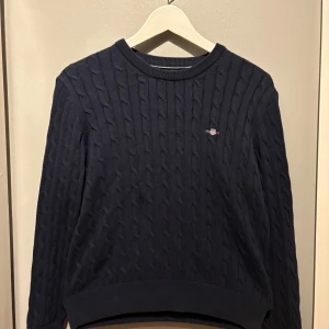 Mörkblå kabelstickad tröja från Gant - Kabelstickad mörkblå tröja från GANT. Tröjan har ribbade muddar vid ärmslut och nederkant, och är tillverkad i mjuk bomull. Perfekt för dig som gillar stickade tröjor. Nypris: 999kr