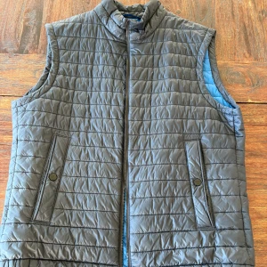 Grå quiltad dunväst från BOSS - Quiltad blå dunväst från BOSS med dragkedja framtill och två fickor med knappstängning. Insidan är blå och västen har hög krage. Tillverkad i syntetmaterial med Primaloft-isolering för extra värme. Perfekt för lager-på-lager-stil.