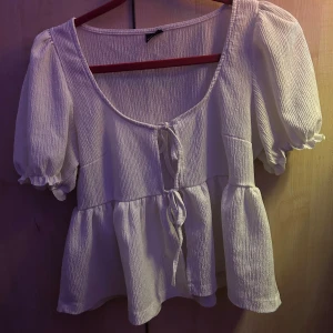 Vit blus med knyt från Gina Tricot - Supersöt vit blus från Gina Tricot i storlek S. Den har puffiga korta ärmar, volang nedtill och två knyt framtill. Tyget är lätt strukturerat och ger en luftig känsla. Perfekt för dig som gillar romantiska och trendiga plagg. Bra skick.