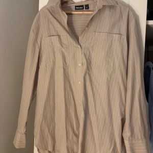 Beige randig skjorta från Pieces - Snygg beige skjorta från Pieces med tunna vita ränder. Skjortan har klassisk krage, knäppning framtill och två bröstfickor. Långärmad och i luftig bomull, perfekt för en chill och stilren look.