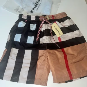 Burberry badbyxor - Snygga badshorts från Burberry med klassiskt rutigt mönster i svart, beige, vitt och rött. Shortsen har elastisk midja och cool snörning i rött, vitt och svart. Perfekta för sommaren och ger en lyxig vibe vid poolen eller stranden.
