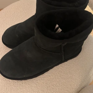 Svarta UGG boots i mocka - Klassiska svarta UGG boots i mjuk mocka med rund tå och platt sula. Fodrade med fluffigt material på insidan för extra värme och komfort. Perfekta för kyliga dagar och enkel att matcha till olika outfits.