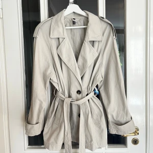 Kort trenchcoat med knytband 100% bomull - Trendig kort beige trenchcoat med knytband i storlek S. Materialet är 100% bomull, i fint skick! Mycket elegant, extra knappar medföljer