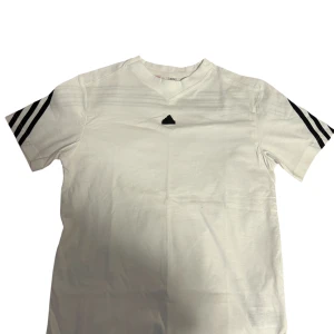 Vit Adidas t-shirt med svarta ränder - Vit t-shirt från Adidas med klassiska svarta ränder på axlar och ärmar. T-shirten har en enkel v-ringning och Adidas-logga framtill. Tillverkad i mjuk bomull och har en normal passform. Perfekt för en sportig och clean look. Till pojkar 13-14 år 
