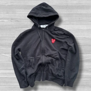 Comme des Garçons Play  - Svart hoodie från Comme des Garçons Play med dragkedja och huva. Tröjan har det ikoniska röda hjärta-loggan med ögon broderad på bröstet. Tillverkad i mjuk bomull och har fickor framtill. Perfekt för dig som gillar streetwear och stilrena detaljer.