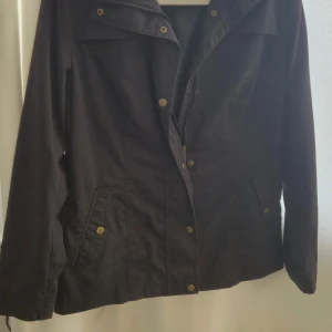 Svart vindjacka från Bonprix, stl 38 - Svart vindjacka från Bonprix Collection med dragkedja och tryckknappar framtill. Jackan har två fickor med lock och guldiga knappar, samt klassisk krage. Perfekt för dig som vill ha en enkel och stilren jacka till vardags.