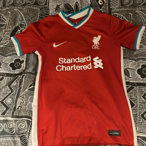 Äkta Liverpool Nike röd matchtröja  - Liverpool FC matchtröja från Nike i rött med vita och turkosa detaljer vid krage och ärmslut. Tröjan har klubbmärke och sponsortryck på bröstet samt Nike Dri-FIT-material som andas. Klassisk V-ringning och korta ärmar, perfekt för fotbollsfans. Tröjan är äkta och i bra skick. Storleken är m i barn och det står 134-144 cm.