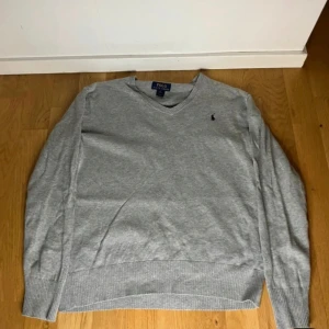 Grå v-ringad tröja från Polo Ralph Lauren - Klassisk grå v-ringad tröja från Polo Ralph Lauren med diskret broderad logga på bröstet. Tröjan har långa ärmar och ribbade muddar vid ärmslut och nederkant. Perfekt för lager-på-lager och stilren look.