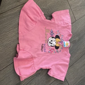 Rosa Minnie Mouse topp från H&M - Supersöt rosa topp med volangkant och korta ärmar från H&M. Framsidan har ett färgglatt tryck av Minnie Mouse med blommor och gul rosett. Perfekt för små Disney-fans som vill sticka ut lite extra.