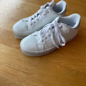 Vita sneakers med glittrig sula - Snygga vita sneakers med glittriga detaljer på sulan och hälen. Skorna har klassisk snörning, rund tå och är tillverkade i syntetmaterial. Perfekta för dig som vill sticka ut lite extra med dina skor. Skorna är endast använda en gång. Priset kan diskuteras.