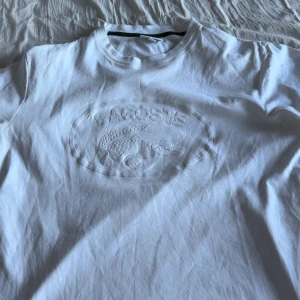 Vit t-shirt från Lacoste med logga - Vit t-shirt från Lacoste med diskret, upphöjd logga och krokodil på bröstet. Klassisk rund halsringning och korta ärmar. Tillverkad i mjuk bomull för en clean och fräsch look. Liten grön krokodil på ryggen vid nacken.