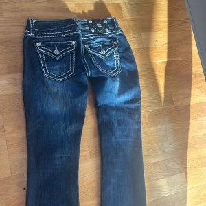 Miss Me jeans med vita sömmar - Mörkblå jeans från Miss Me med kontrasterande vita sömmar och snygga detaljer på bakfickorna. Byxorna har dekorativa knappar och broderier, samt en klassisk bootcut-modell. Materialet är jeans och passformen är normal. Perfekta för dig som gillar statement-jeans.