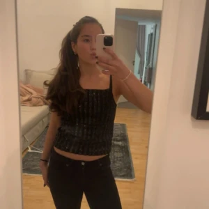 Svart glittrig linne-topp - Säljer en svart glittrig linne-topp med smala axelband och croppad passform. Toppen har ett snyggt paljettmönster som ger extra bling och är perfekt för att sticka ut. Enkel och stilren design som passar till många olika looks.