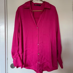 Rosa skjorta/blus från Zara - Säljer en snygg skrikrosa skjorta från Zara med krage och knappar framtill. Skjortan har lång ärm med manschett och flowy passform. Mjukt, glansigt material som ger en lyxig känsla. Generös passform, passar S-M och kanske även L beronde på armstorlek. Använd 2 gånger. 