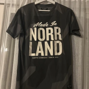 Svart t-shirt från SQRTN Company - Cool svart t-shirt från SQRTN Company med stort vitt tryck 'Made in Norrland' på bröstet. T-shirten har korta ärmar och en klassisk passform. Materialet är mjuk bomull och den har diskreta geometriska mönster i grått.