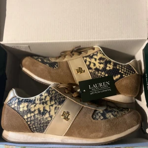 Sneakers från Lauren Ralph Lauren - Snygga sneakers från Lauren Ralph Lauren med beige och bruna detaljer i mocka och skinn. Skorna har snörning och RL-logga i guld på sidan. Perfekta för dig som vill sticka ut med en unik stil.