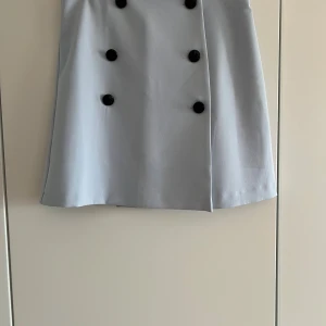 Ljusblå kortkjol med svarta knappar - Snygg ljusblå kortkjol med sex svarta dekorativa knappar framtill. Kjolen har en rak passform och är perfekt för dig som gillar stilrena detaljer. Enkel att matcha med både t-shirt och blus.