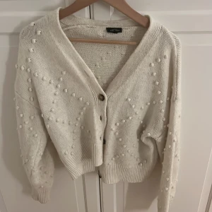 Vit stickad kofta från KappAhl - Superfin vit stickad kofta från KappAhl i storlek S. Koftan har dekorativa bollar i stickningen, v-ringning och knappar framtill.