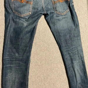 Blå Nudie Jeans W28 L32 - Säljer ett par klassiska blå jeans från Nudie Jeans med snygga slitningar och orange kontrastsömmar. Modellen har fem fickor, normal passform och raka ben. Jeansen är tillverkade i bomull och har Nudie-logga på bakfickan. Pris kan diskuteras.